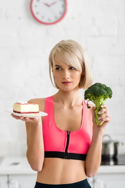 Chica rubia sosteniendo brócoli verde y platillo con pastel dulce en las manos - foto de stock