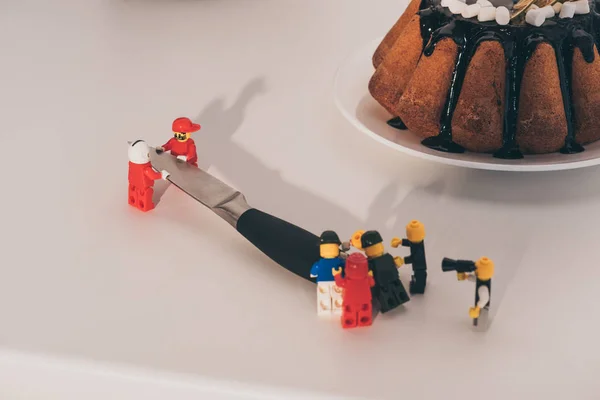 KYIV, UKRAINE - MARCH 15, 2019: plastic lego figurines carrying metal knife near cake on white — Stock Photo