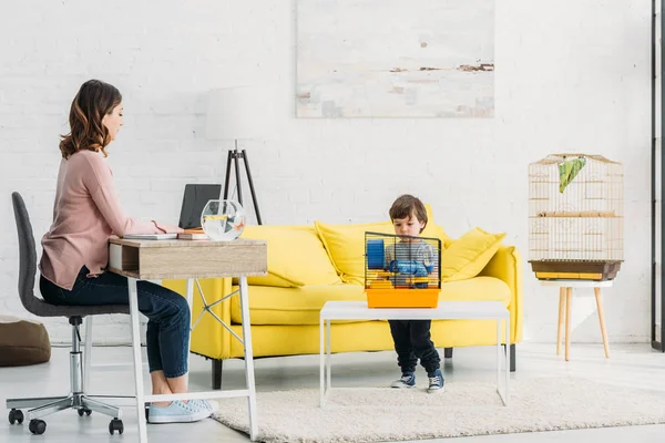 Attractive mother using laptop and cute son standing near pet cage — Stock Photo