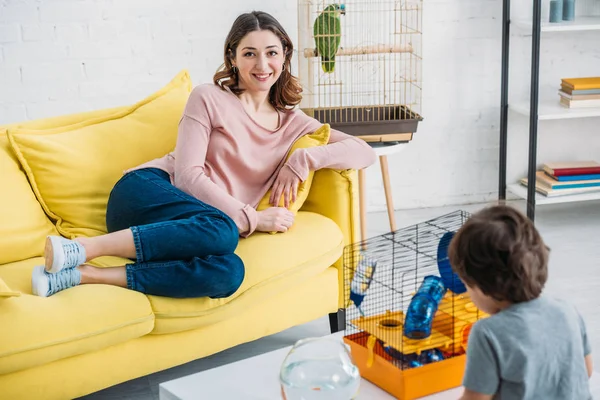 Foyer sélectif à la mère souriante reposant sur un canapé jaune tandis que le fils mignon debout près de la table avec cage pour animaux de compagnie — Photo de stock