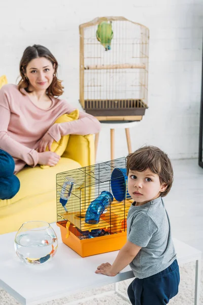 Messa a fuoco selettiva del bambino carino in piedi vicino al tavolo con ciotola di pesce e gabbia per animali domestici, e madre sorridente appoggiata sul divano giallo — Foto stock