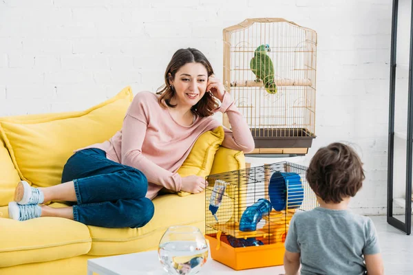 Улыбающаяся мать отдыхает на желтом диване рядом с птичьей клеткой с попугаем, а сын стоит за столом с домашним животным и аквариумом — стоковое фото