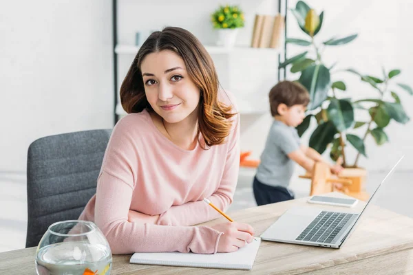 Mulher atraente sentado à mesa perto do laptop, sorrindo e olhando para a câmera — Fotografia de Stock