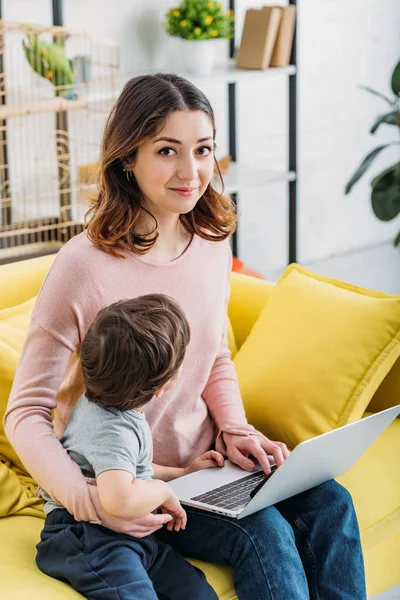 Bella donna con figlio carino utilizzando il computer portatile mentre seduto sul divano giallo a casa — Foto stock