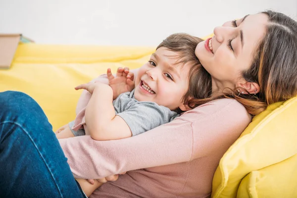 Feliz madre con hijo alegre descansando en el sofá amarillo en casa - foto de stock
