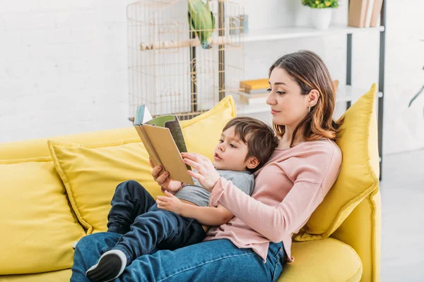 Femme souriante avec livre de lecture mignon enfant sur canapé ensemble — Photo de stock