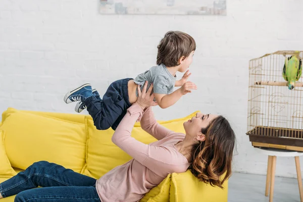 Glückliche junge Mutter mit entzückendem Jungen hat Spaß auf gelbem Sofa in der Nähe von Vogelkäfig mit grünem Papagei — Stockfoto