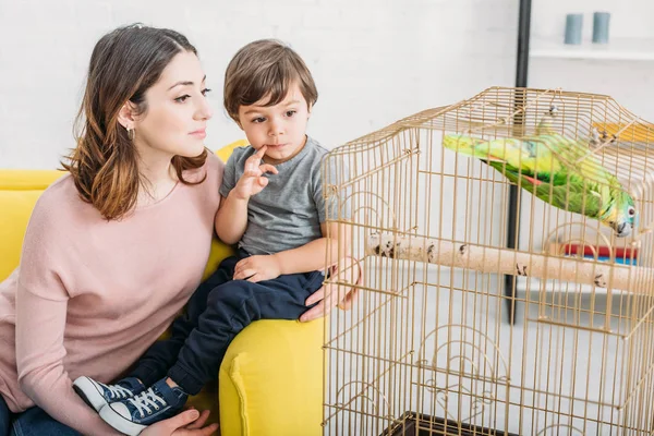 Hübsche Frau mit süßem Jungen auf Sofa sitzend und grünen Papagei im Vogelkäfig zu Hause anschauend — Stockfoto