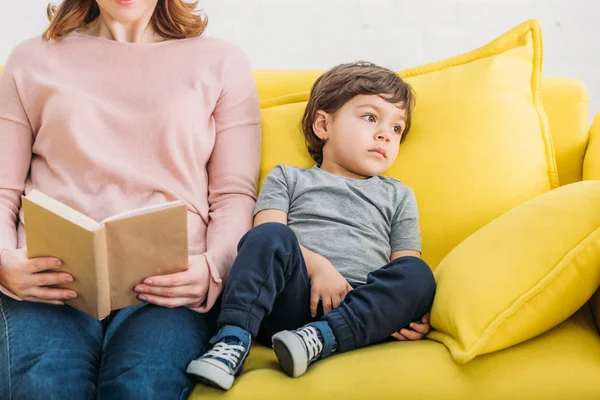 Teilansicht einer Frau, die Buch in der Nähe ihres entzückenden Sohnes liest, der zu Hause auf dem Sofa liegt — Stockfoto