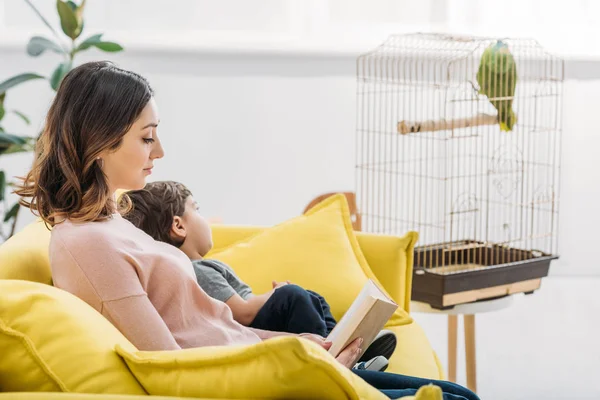 Atractiva mujer con libro y lindo chico descansando en sofá cerca de jaula de pájaro - foto de stock