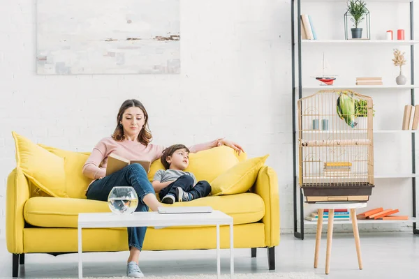 Séduisante femme avec adorable fils reposant sur un canapé jaune dans un salon spacieux — Photo de stock