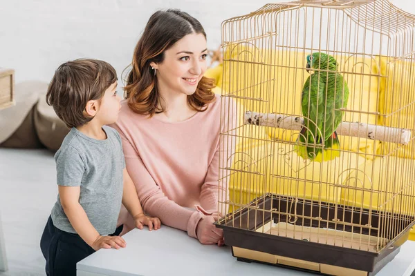 Smiling attractive woman with adorable son looking at green parrot in bird cage — Stock Photo