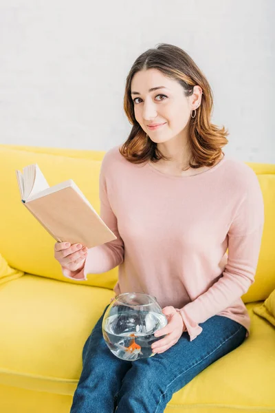Hübsche Frau mit Fischschale und Buch auf gelbem Sofa sitzend und in die Kamera blickend — Stockfoto