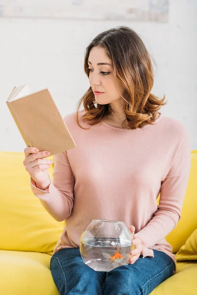 Attractive young woman reading book while holding fish bowl on knees — Stock Photo