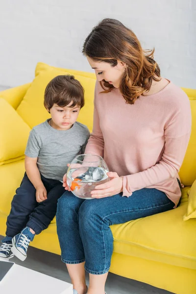 Sonriente madre sosteniendo pecera mientras está sentada en un sofá amarillo con su adorable hijo - foto de stock