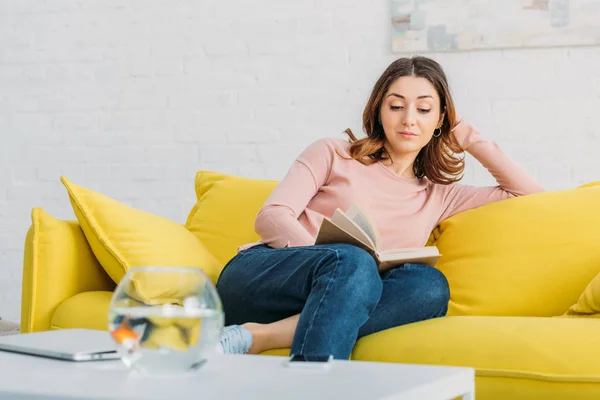 Schöne Frau liest Buch, während sie sich zu Hause auf dem gelben Sofa ausruht — Stockfoto