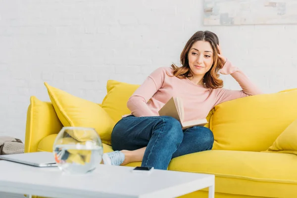 Красива жінка з книгою відпочиває на жовтому дивані вдома — стокове фото