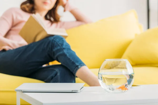 Cropped view of woman resting with book on sofa near table with fish bowl — Stock Photo