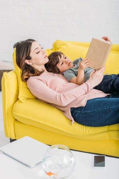 Jovem mãe com adorável filho leitura livro enquanto deitado no sofá juntos — Fotografia de Stock