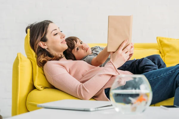 Pretty smiling woman  with cute boy reading book while lying on sofa together — Stock Photo
