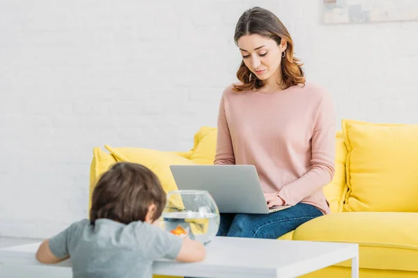 Mujer bonita usando el ordenador portátil, mientras que el hijo adorable mirando pecera - foto de stock