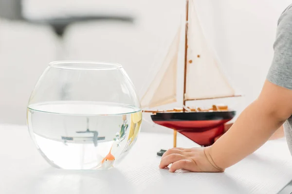 Vue partielle du garçon debout près du bol à poisson et du modèle de bateau en bois — Photo de stock