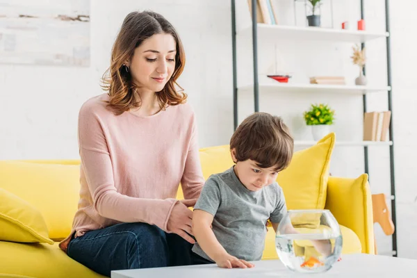 Menino curioso bonito olhando para o aquário com peixe dourado perto de mãe sorridente — Fotografia de Stock