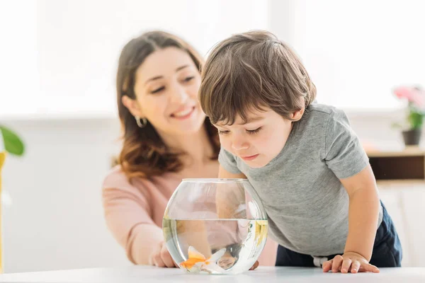 Selective focus of adorable child looking into fish bowl near smiling mother — Stock Photo