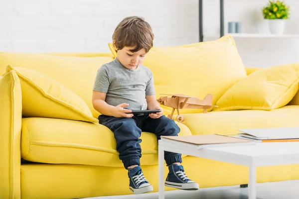 Menino bonito sentado no sofá amarelo perto de modelo de avião de madeira e segurando tablet digital — Fotografia de Stock