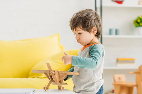 Netter aufmerksamer Junge spielt zu Hause mit hölzernem Flugzeugmodell — Stockfoto