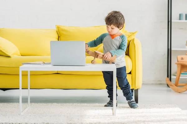 Adorable niño jugando con el modelo plano de madera cerca del ordenador portátil en casa - foto de stock