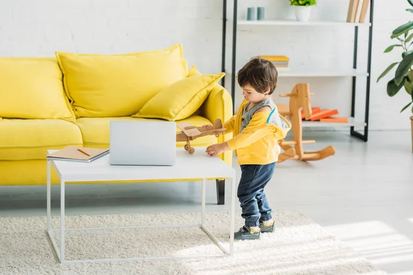 Carino ragazzo giocando con modello aereo di legno vicino tavolo con computer portatile a casa — Foto stock