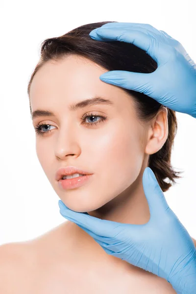 Обрізаний вид пластичного хірурга в синіх латексних рукавичках, що торкаються обличчя красивої жінки, ізольованої на білому — стокове фото