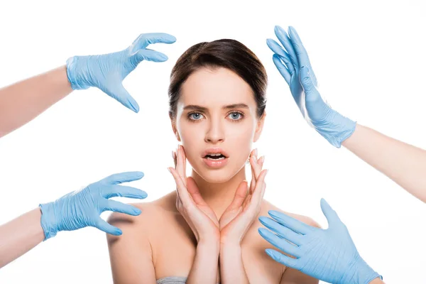 Обрезанный вид пластических хирургов в синих латексных перчатках возле лица удивленной девушки, изолированной на белом — стоковое фото