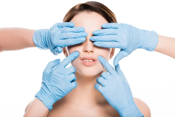 Vista recortada de cirujanos plásticos en guantes de látex azul cubriendo los ojos y tocando la cara de niña aislada en blanco - foto de stock