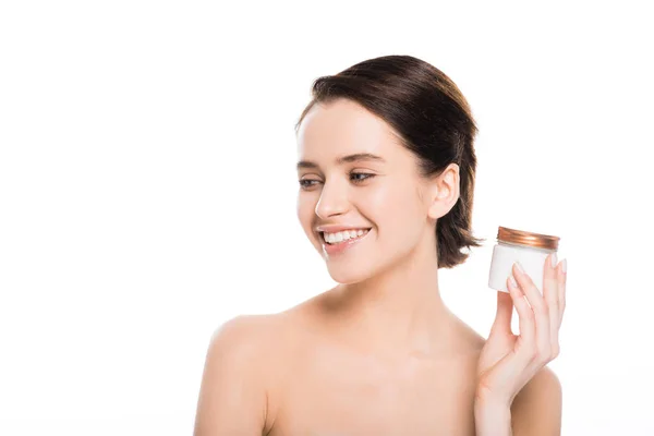 Mujer joven desnuda y morena sosteniendo contenedor con crema facial y sonriendo aislado en blanco - foto de stock