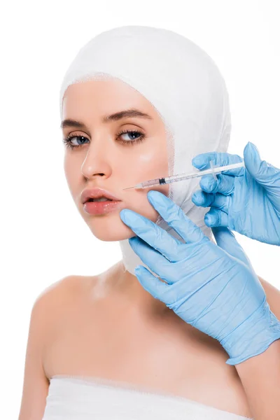 Cropped view of cosmetologist in latex gloves holding syringe near face of girl with bandaged head isolated on white — Stock Photo