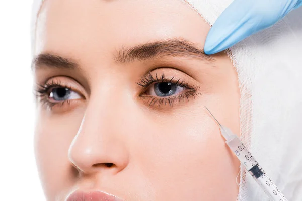 Vista recortada del cosmetólogo en guantes de látex que sostienen la jeringa cerca del ojo de la mujer con la cabeza vendada aislada en blanco - foto de stock