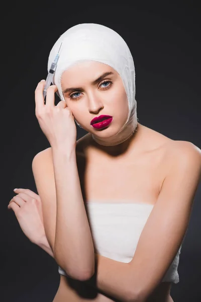Schöne Frau mit bandagiertem Kopf hält Spritze isoliert auf grau — Stockfoto