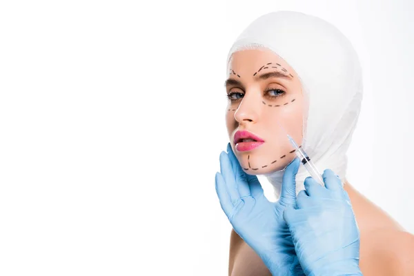 Обрезанный вид пластического хирурга в латексных перчатках, держащего шприц возле лица молодой женщины с отметинами, изолированными на белом — стоковое фото