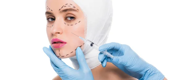 Панорамный снимок пластического хирурга в латексных перчатках, держащего шприц возле лица молодой женщины с отметинами, изолированными на белом — стоковое фото