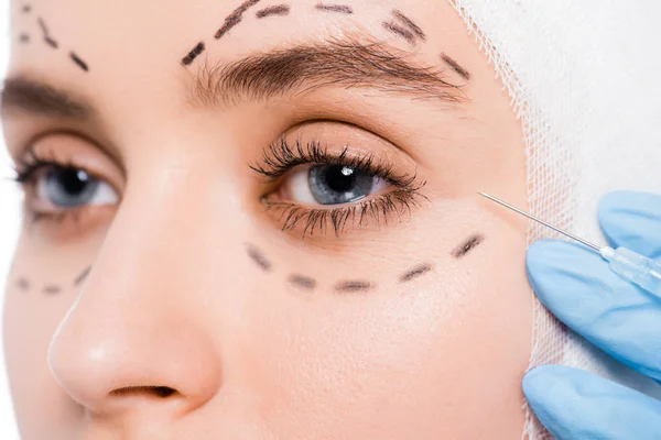 Vista recortada del cirujano plástico en guante de látex que sostiene la jeringa cerca de la cara de la mujer con marcas aisladas en blanco - foto de stock
