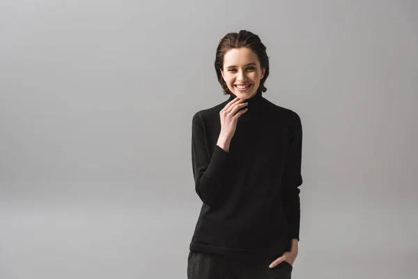Весела молода жінка в чорному джемпері стоїть з рукою в кишені на сірому — стокове фото