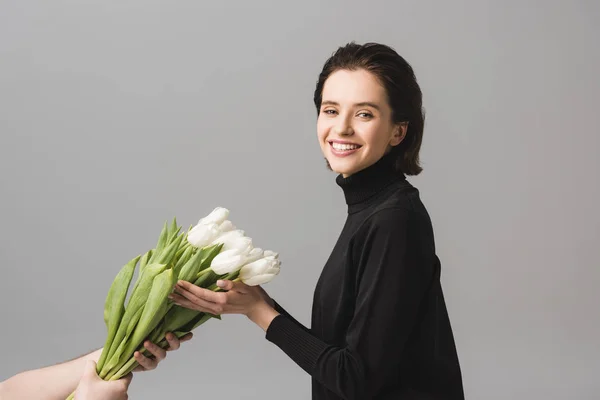 Vista recortada del hombre sosteniendo tulipanes blancos cerca de la joven feliz aislado en gris - foto de stock