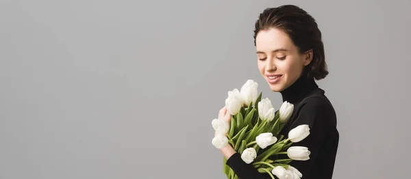 Panoramaaufnahme einer glücklichen brünetten Frau, die weiße Tulpen isoliert auf grau betrachtet — Stockfoto