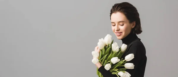 Colpo panoramico di donna bruna felice guardando tulipani bianchi isolati su grigio — Foto stock