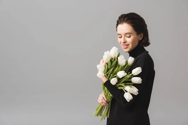 Cheerful brunette young woman looking at white tulips isolated on grey — Stock Photo