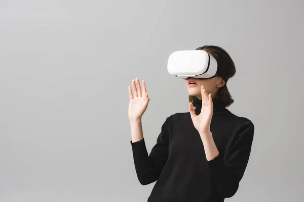 Sorpresa donna bruna indossando cuffie realtà virtuale mentre gesticolava isolato sul grigio — Foto stock