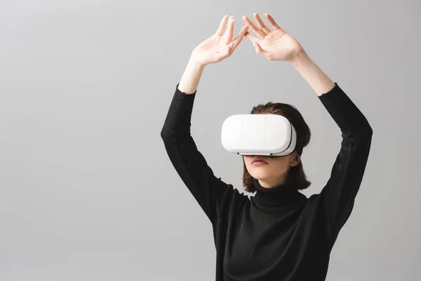 Brunette woman wearing virtual reality headset while standing with hands above head isolated on grey — Stock Photo