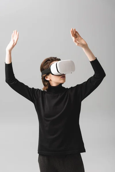 Donna che indossa auricolare realtà virtuale mentre in piedi con le mani sopra la testa isolato su grigio — Stock Photo